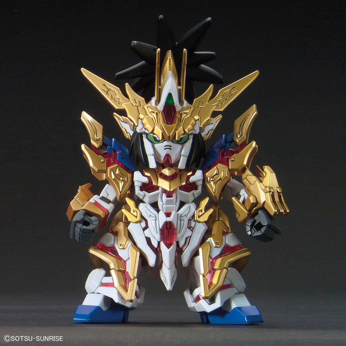 A small-scale collectible model of a Gundam-style mecha in metallic gold, white, blue, and red armor, photographed against a dark gray studio background. A copyright watermark for SOTSU-SUNRISE is visible in the bottom-left corner.