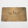 A rectangular brown cardboard mat with rounded corners, featuring a distressed, crumpled cardboard texture and the printed black text 'THIS SIDE UP (FOR WINNING)' with an arrow pointing up in the center, isolated on a white background.