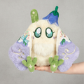 Plush toy with a white face, green hands and feet, and purple wings held by a hand on a white background