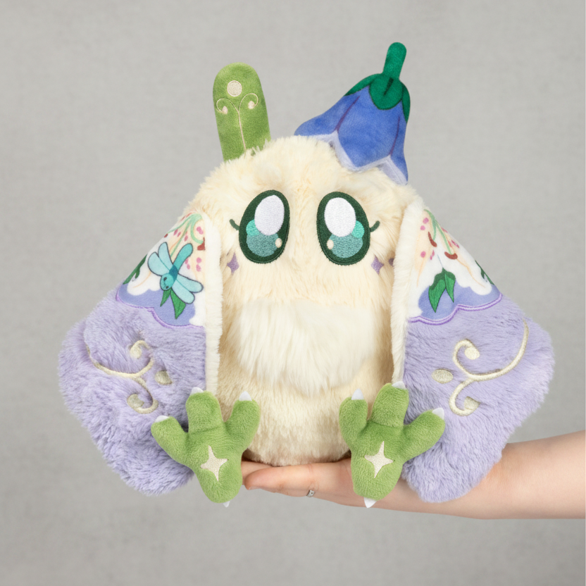 Plush toy with a white face, green hands and feet, and purple wings held by a hand on a white background