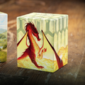 A close-up of a square collectible card or token featuring a red dragon over a yellow landscape, standing on a wooden surface. A second illustrated card is partially visible at the left edge.